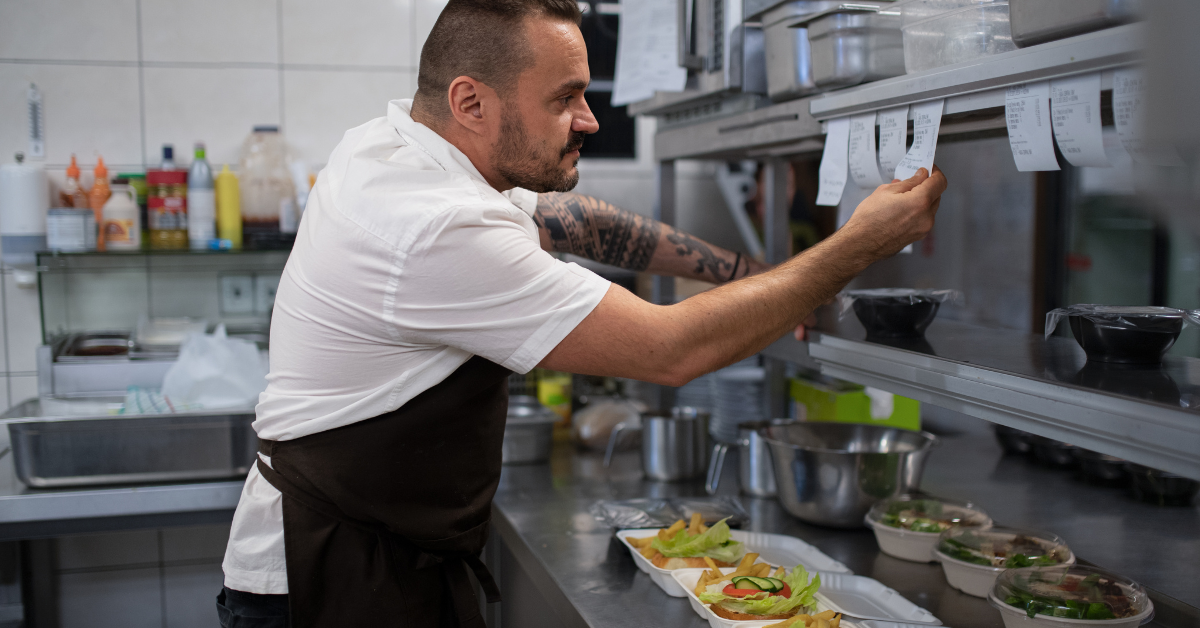 Chef in restaurant kitchen checking paper order tickets above prepared dishes.