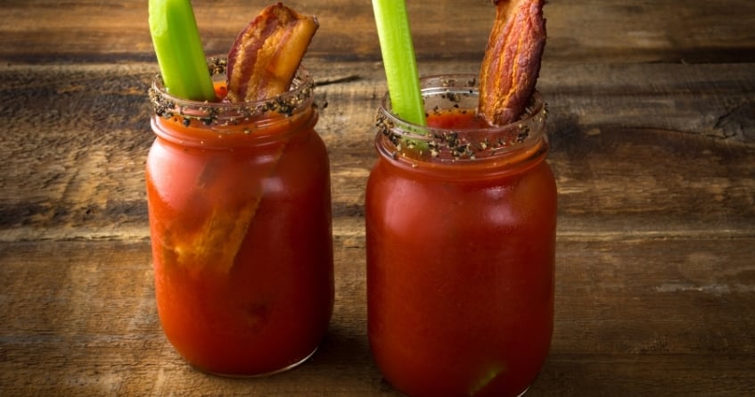 Two Bloody Mary cocktails served in mason jars with peppered rims, celery sticks, and crispy bacon garnish — a rustic, brunch-style presentation popular in casual bars and restaurants.