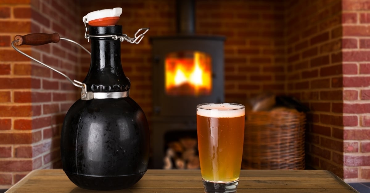 Glass growler and pint of beer placed on a wooden table in front of a fireplace.