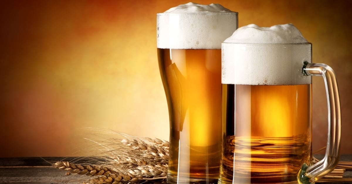 Two frothy pints of draft beer on a wooden bar table with wheat stalks, symbolizing beverage costs and profit margins