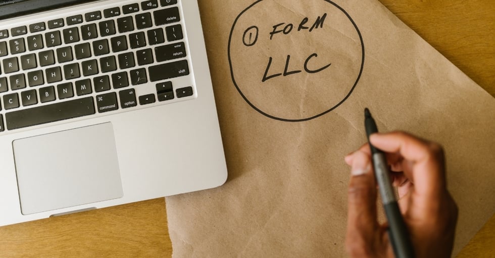 Hand writing “Form LLC” on paper next to a laptop — first step in starting a bar business.