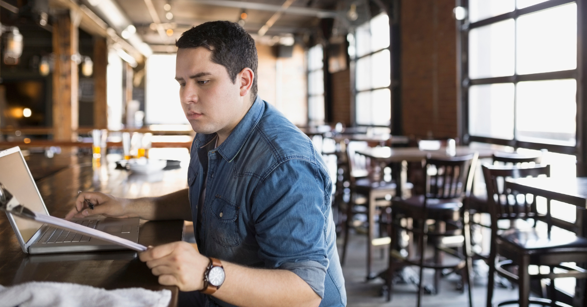 Restaurant manager reviewing invoices on a laptop and clipboard — careful invoice reconciliation ensures inventory records match actual costs.