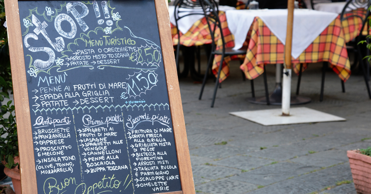 Close-up of a handwritten chalkboard menu outside a casual Italian restaurant with checkered tablecloths in the background.
