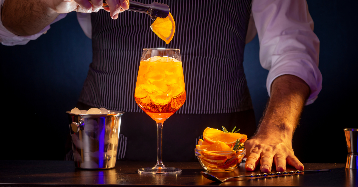 Bartender garnishing a bright orange cocktail with a citrus peel at a dimly lit bar, highlighting attention to detail in drink presentation.