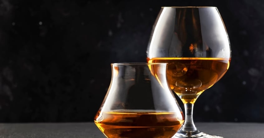Two cognac snifters filled with amber spirits against a dark background, showcasing the rounded bowl and narrow rim that enhance aroma and warmth during service.