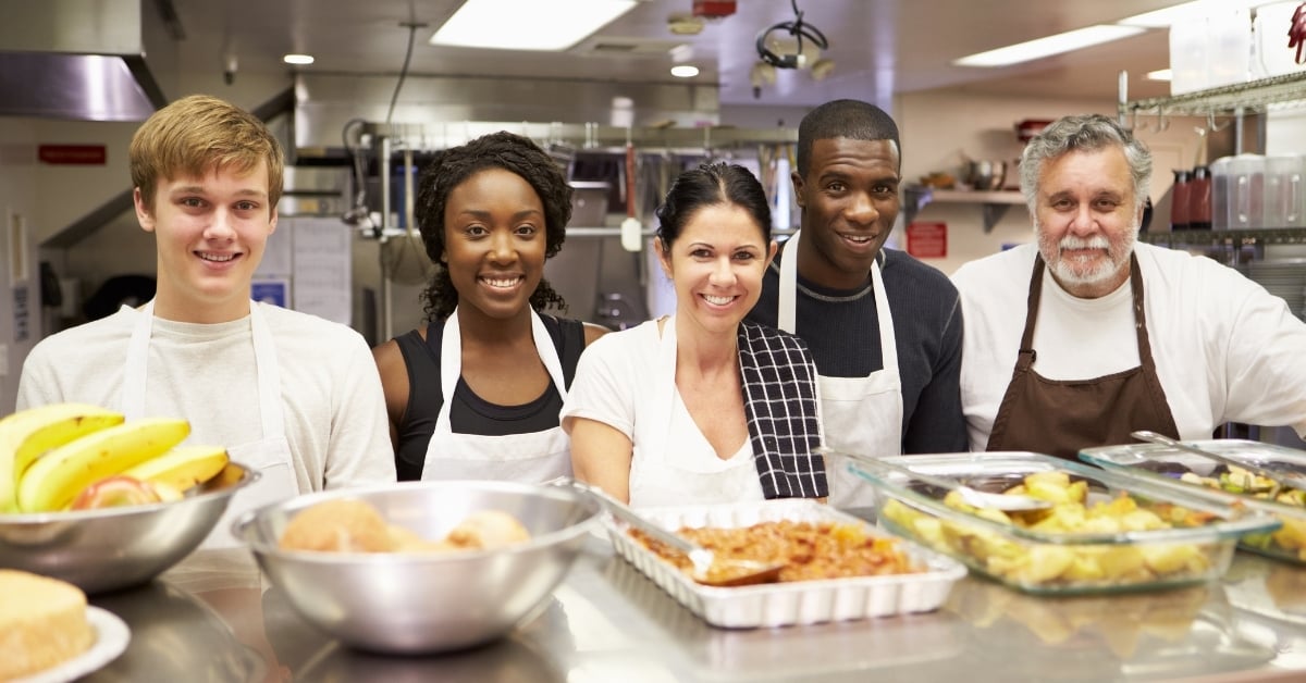 Restaurant kitchen team smiling behind the counter, wearing aprons and standing with prepared food trays and fresh produce.