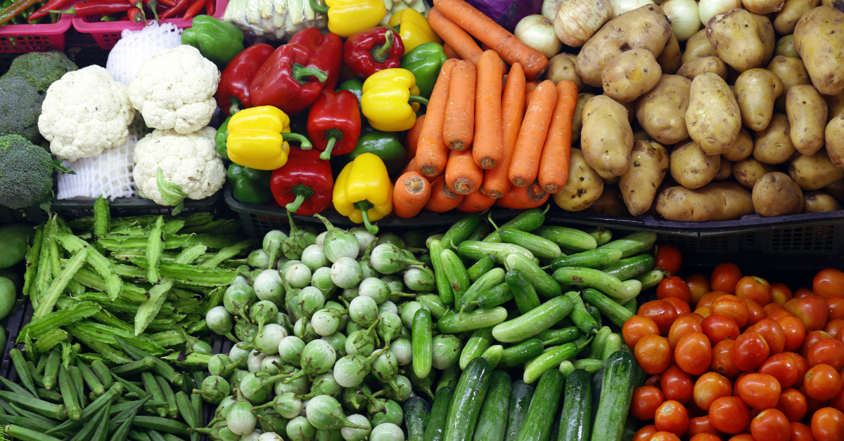 Colorful display of fresh, locally sourced vegetables including carrots, potatoes, tomatoes, bell peppers, cauliflower, and cucumbers at a market or restaurant supplier.