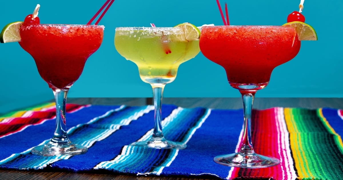 Three colorful margaritas in classic double-bowl glasses with salted rims and fruit garnishes, illustrating variety and portion control in specialty service.