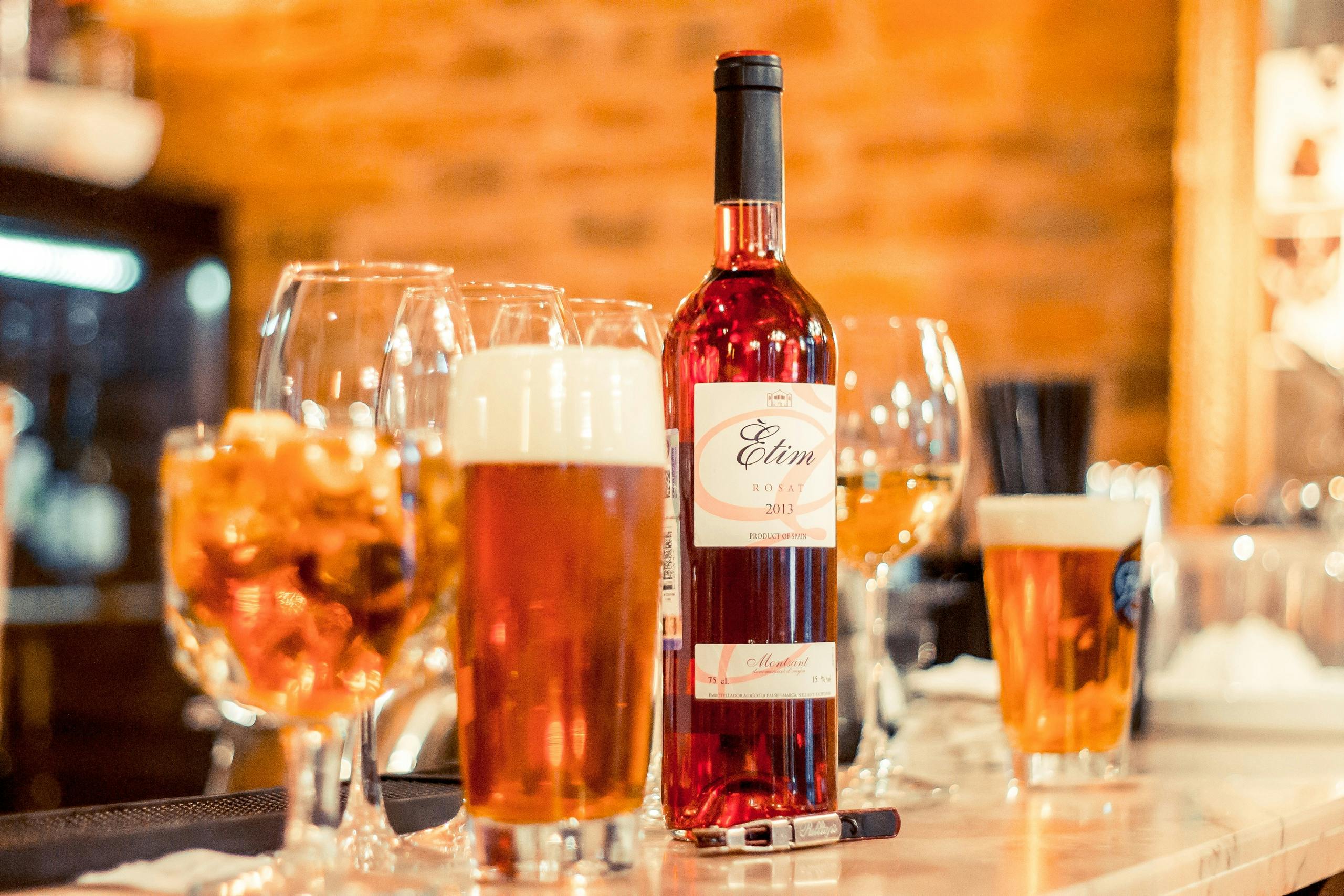 Close-up of a bar counter featuring a glass of beer, a bottle of rosé wine, wine glasses, and a cocktail, set against a warm, blurred background.