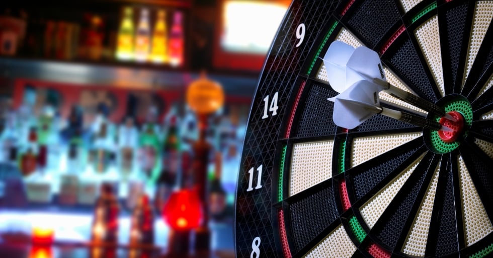 Close-up of three darts hitting the bullseye on an electronic dartboard, with a blurred bar full of liquor bottles in the background—symbolizing precision and bar performance.