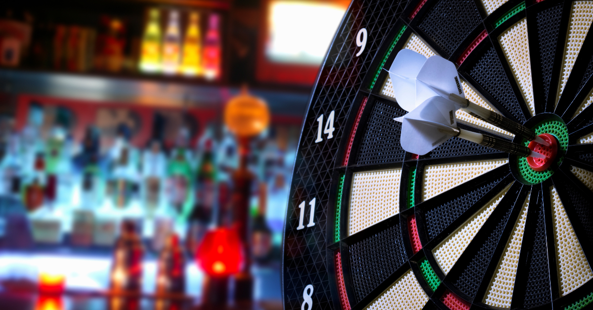 Close-up of three darts hitting the bullseye on an electronic dartboard, with a blurred bar full of liquor bottles in the background—symbolizing precision and bar performance.