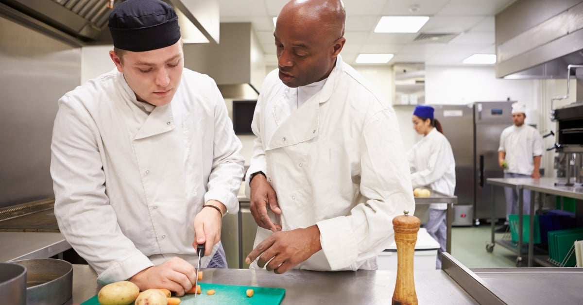 Chef coaching kitchen staff on knife skills and food preparation — illustrating effective restaurant training and operational consistency.