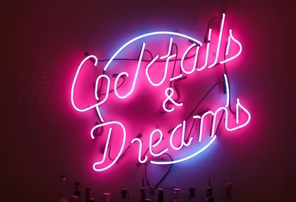 Neon sign reading “Cocktails & Dreams” glowing in pink and blue against a dark bar background.