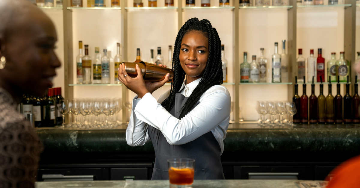 Smiling bartender shaking a cocktail shaker behind a bar, with liquor bottles and glassware neatly displayed on backlit shelves.