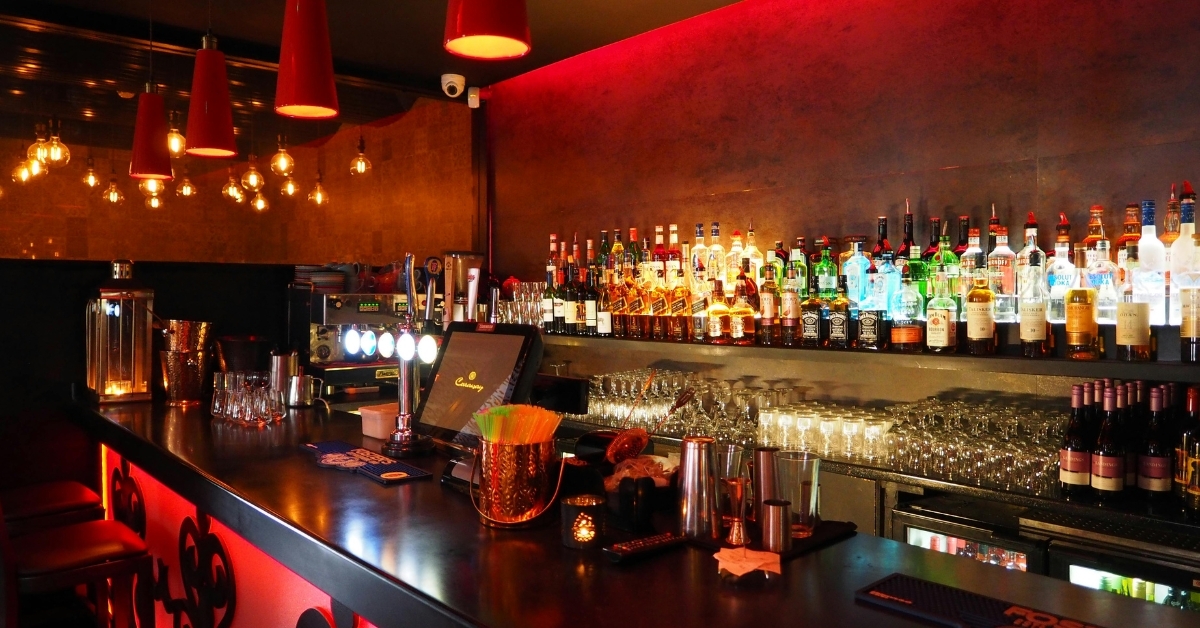Modern bar interior with shelves of liquor bottles, illuminated backlighting, cocktail tools, and pendant lights creating a warm atmosphere.