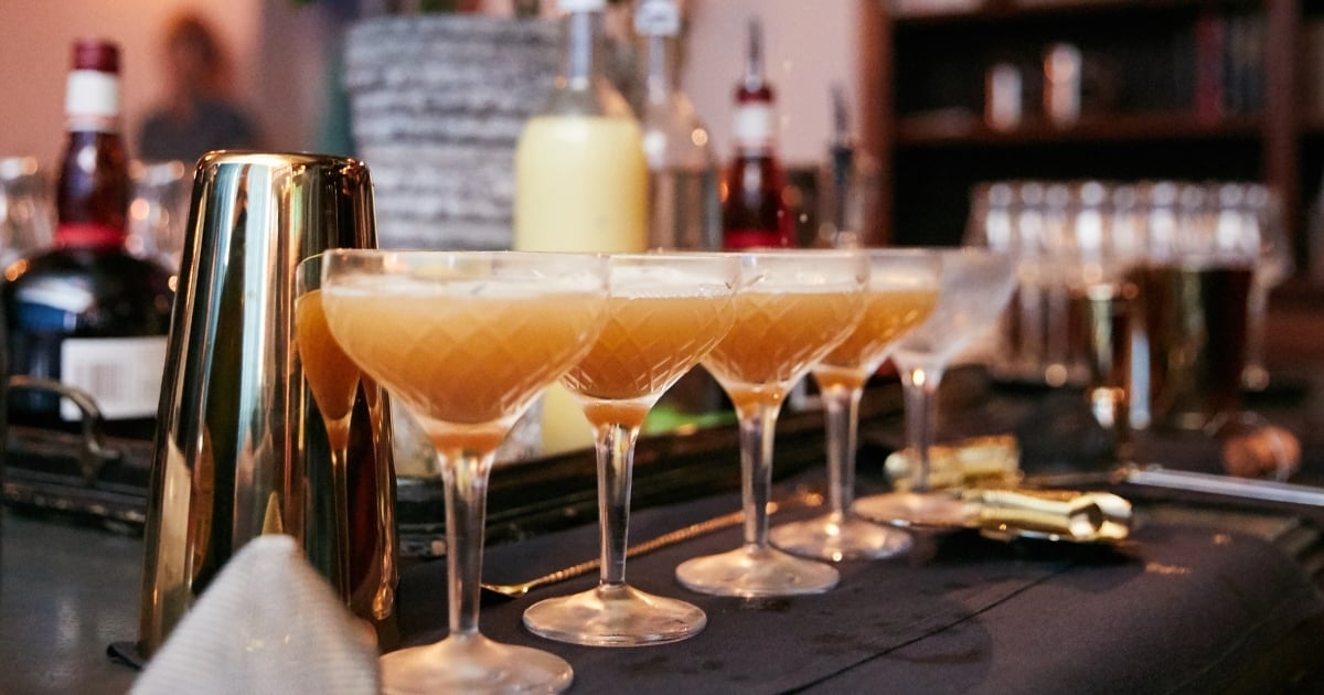 A row of freshly batched cocktails in coupe glasses on a bar, illustrating consistent pour sizes used in a cocktail batching calculator.