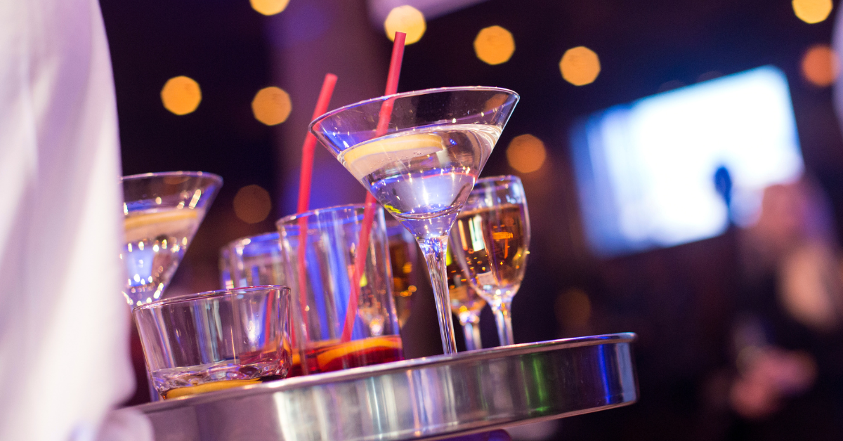 Server carrying a tray of cocktails and martinis in a dimly lit bar during peak hours, showcasing vibrant drinks and fast-paced restaurant service.